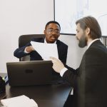 Dois profissionais de terno analisam documentos e gráficos durante uma reunião em um escritório, com um notebook sobre a mesa.