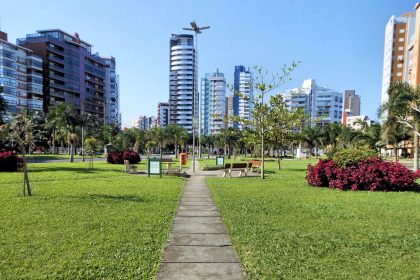 Parque urbano arborizado em Porto Alegre, com edifícios altos ao fundo sob céu azul.
