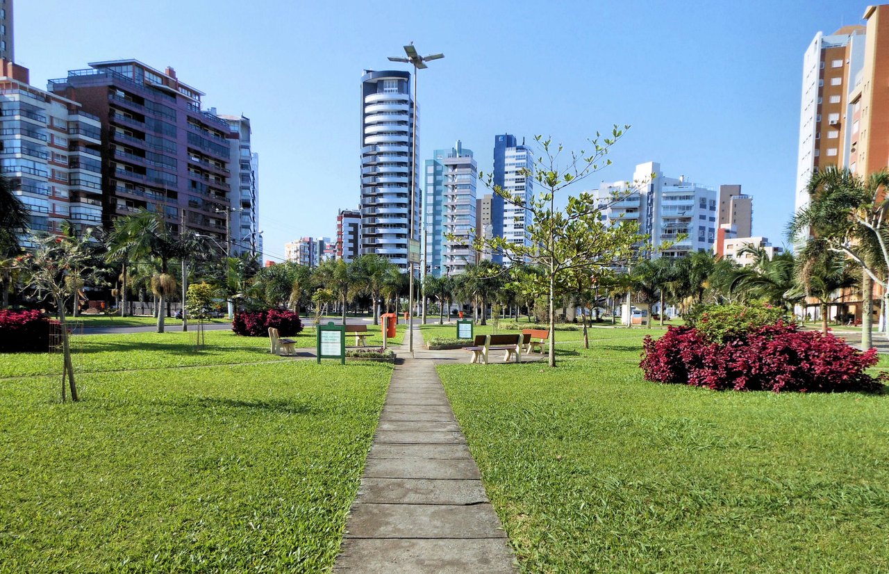 Parque urbano arborizado em Porto Alegre, com edifícios altos ao fundo sob céu azul.