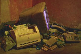 Pilhas de eletrônicos antigos, incluindo um computador e uma impressora, em um ambiente de armazenamento.