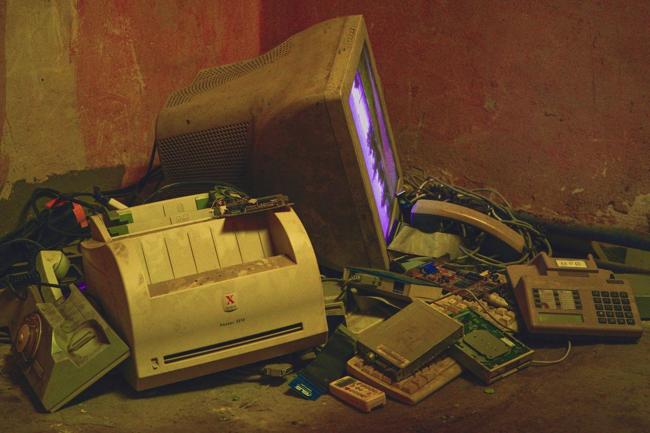 Pilhas de eletrônicos antigos, incluindo um computador e uma impressora, em um ambiente de armazenamento.